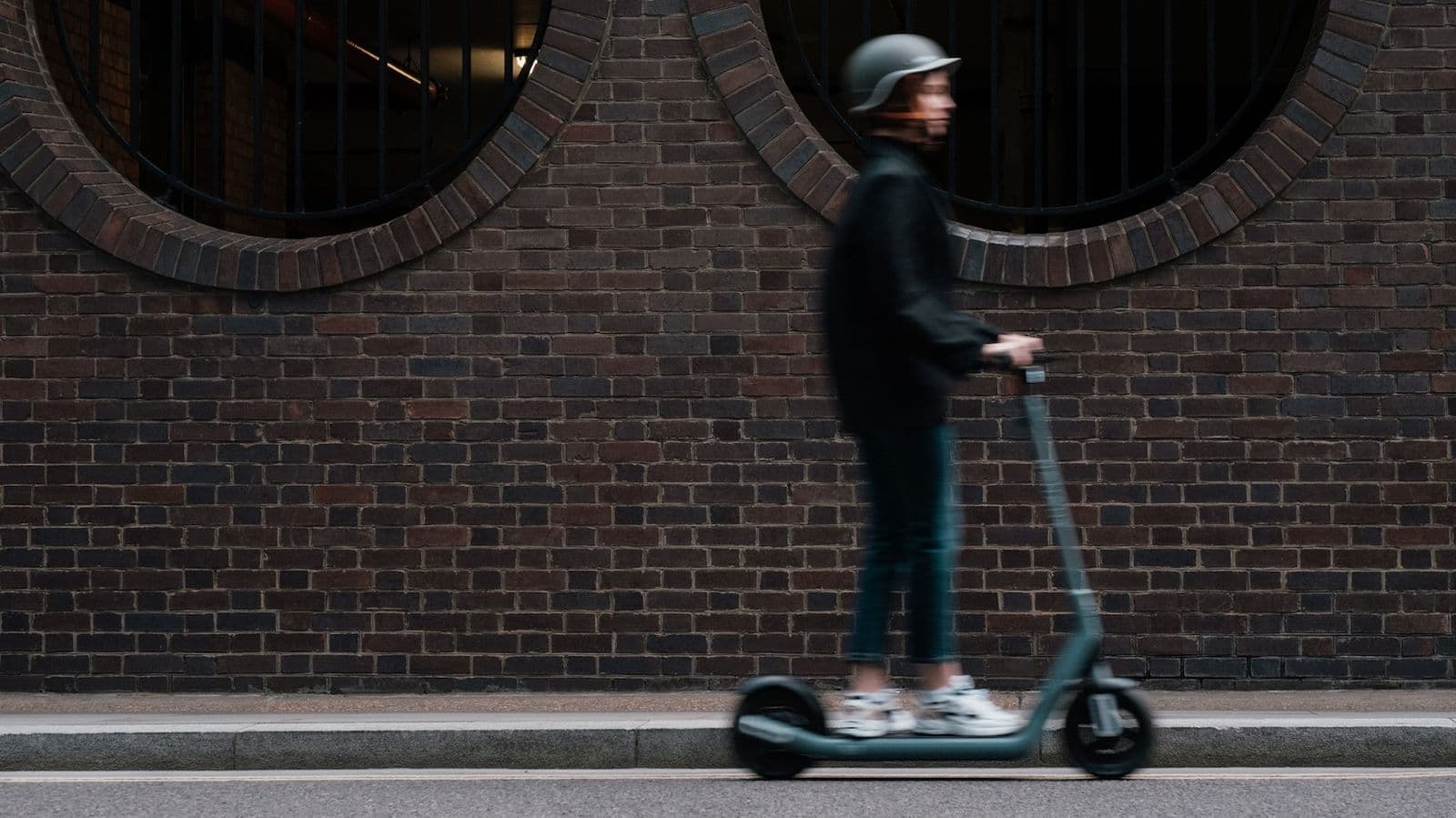 Young woman wearing a black jacket and jeans riding a gray electric scooter from a low angle.